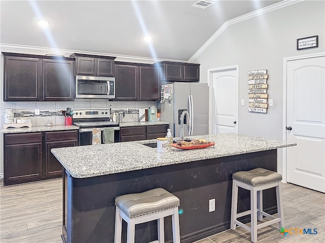 kitchen with an island with sink, vaulted ceiling, light wood-type flooring, and appliances with stainless steel finishes