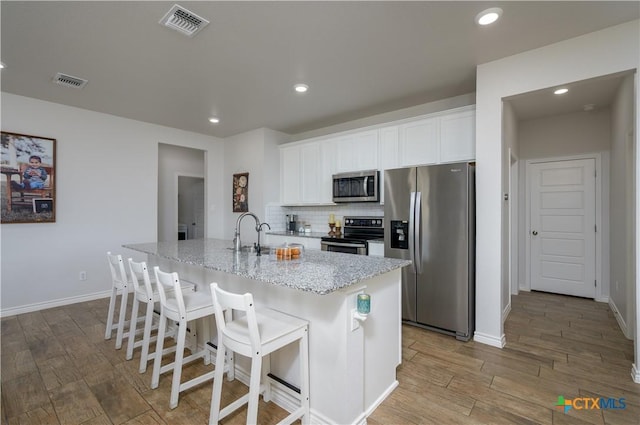 kitchen with light stone counters, a kitchen island with sink, a sink, white cabinetry, and appliances with stainless steel finishes