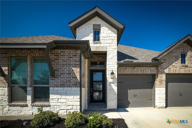 property entrance with stone siding, roof with shingles, concrete driveway, a garage, and brick siding