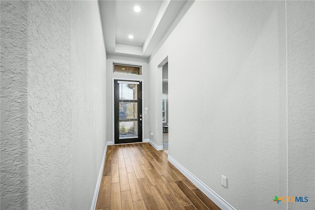 corridor featuring wood finished floors, baseboards, recessed lighting, a raised ceiling, and a textured wall