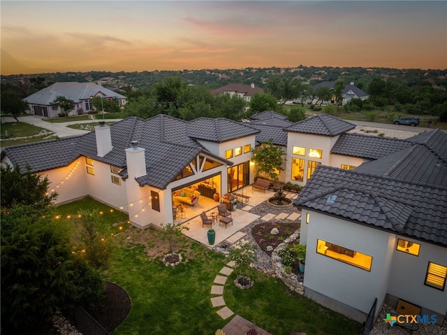 back of property at dusk featuring stucco siding, a chimney, a lawn, a patio, and a tiled roof