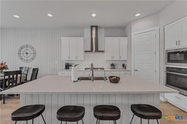 kitchen featuring sink, oven, built in microwave, wall chimney exhaust hood, and an island with sink