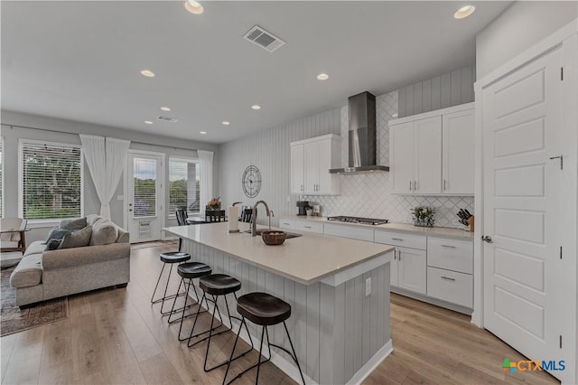 kitchen with an island with sink, stainless steel gas cooktop, a breakfast bar, white cabinets, and wall chimney range hood