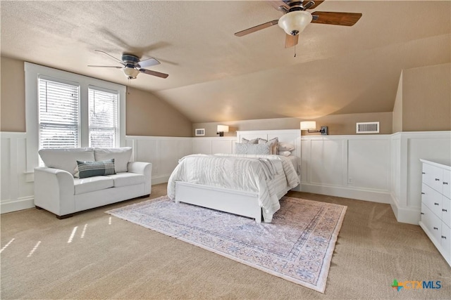 bedroom with a ceiling fan, wainscoting, light colored carpet, vaulted ceiling, and a textured ceiling