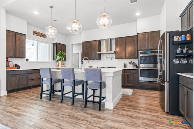 kitchen with dark brown cabinetry, wall chimney exhaust hood, stainless steel appliances, and light countertops