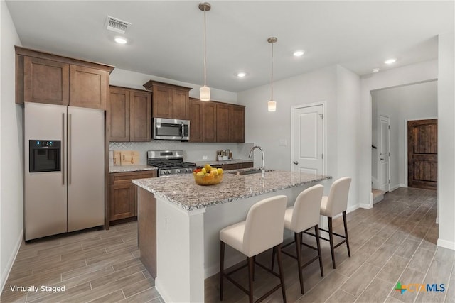 kitchen featuring stainless steel appliances, a kitchen island with sink, pendant lighting, light stone counters, and sink