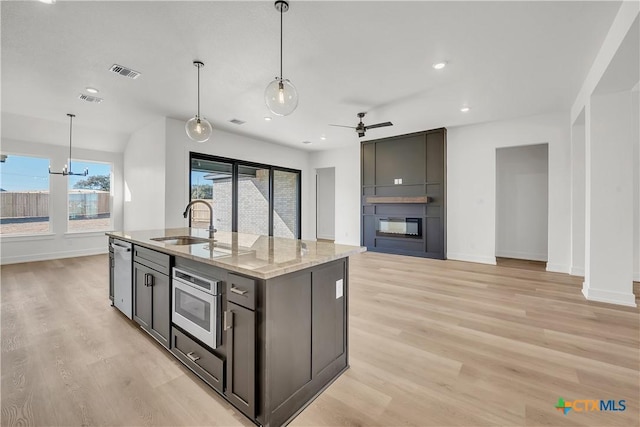 kitchen featuring a fireplace, an island with sink, sink, hanging light fixtures, and light stone counters