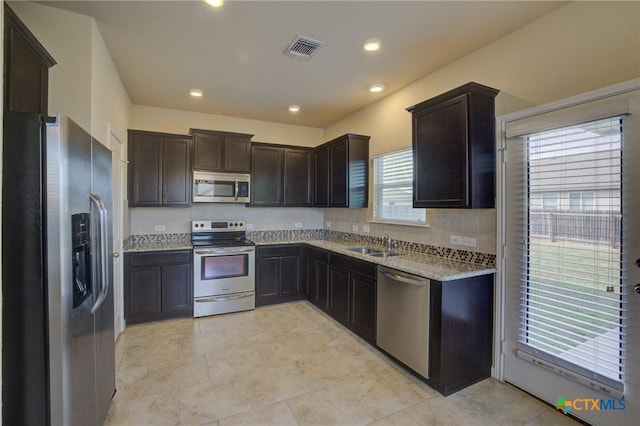 kitchen with appliances with stainless steel finishes, sink, dark brown cabinets, backsplash, and light stone counters