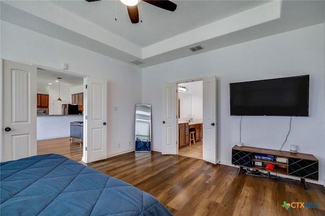 bedroom featuring wood-type flooring, ensuite bath, a tray ceiling, ceiling fan, and stainless steel refrigerator