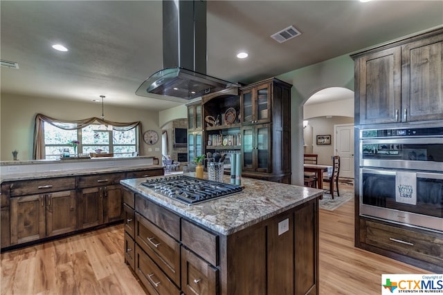 kitchen featuring light wood-type flooring, light stone counters, stainless steel appliances, island range hood, and a center island