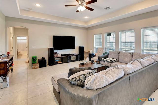 living area featuring a ceiling fan, visible vents, a tray ceiling, light tile patterned flooring, and arched walkways