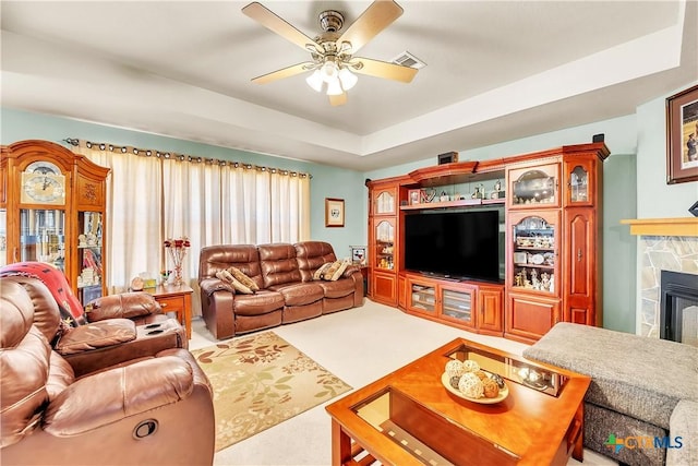 living area featuring carpet, visible vents, a tray ceiling, ceiling fan, and a stone fireplace