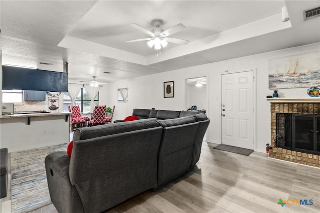 living room featuring ceiling fan, a textured ceiling, a tray ceiling, a fireplace, and light wood-type flooring