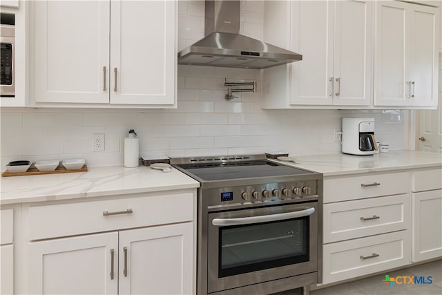 kitchen featuring tasteful backsplash, light stone countertops, white cabinets, wall chimney range hood, and stainless steel range