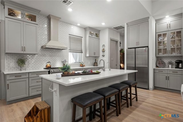 kitchen with wall chimney range hood, a breakfast bar area, appliances with stainless steel finishes, an island with sink, and light wood-type flooring