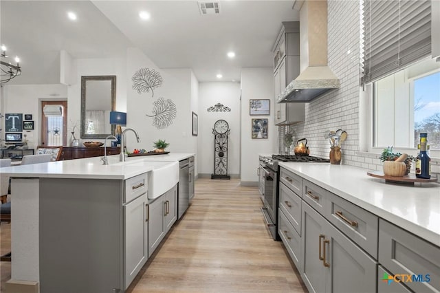 kitchen with wall chimney exhaust hood, sink, light wood-type flooring, stainless steel appliances, and a kitchen island with sink