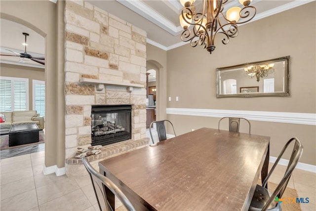 dining space featuring ornamental molding, ceiling fan with notable chandelier, a raised ceiling, a stone fireplace, and light tile patterned flooring