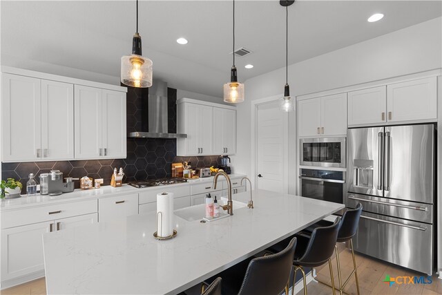 kitchen featuring wall chimney exhaust hood, stainless steel appliances, light stone counters, backsplash, and a center island with sink