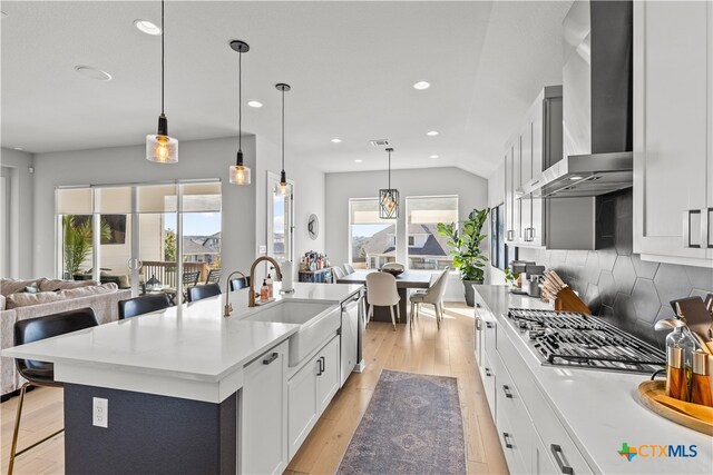 kitchen with light wood-type flooring, sink, wall chimney range hood, pendant lighting, and a center island with sink