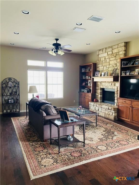 living room featuring a stone fireplace, ceiling fan, visible vents, baseboards, and dark wood-style floors