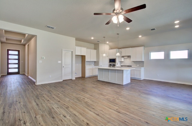 kitchen featuring white cabinets, decorative light fixtures, an island with sink, decorative backsplash, and stainless steel microwave