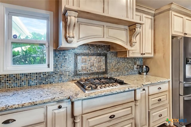 kitchen with appliances with stainless steel finishes, cream cabinetry, light stone counters, and tasteful backsplash