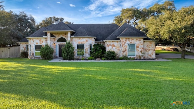 view of front facade with stone siding, roof with shingles, a front yard, and fence