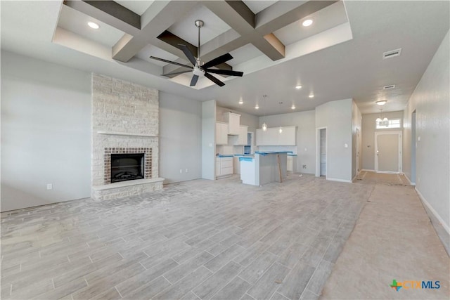 unfurnished living room with light wood finished floors, visible vents, a stone fireplace, coffered ceiling, and ceiling fan with notable chandelier