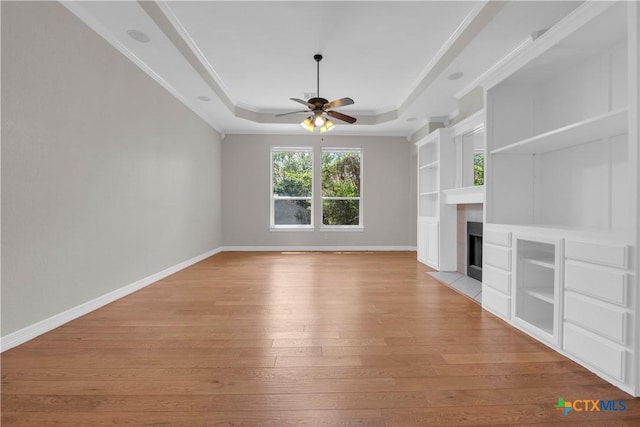 unfurnished living room featuring ceiling fan, a raised ceiling, built in features, ornamental molding, and light wood-type flooring