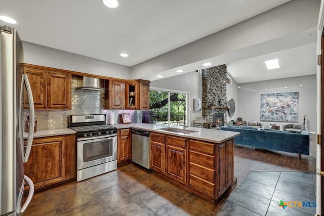 kitchen featuring a stone fireplace, wall chimney range hood, appliances with stainless steel finishes, sink, and kitchen peninsula