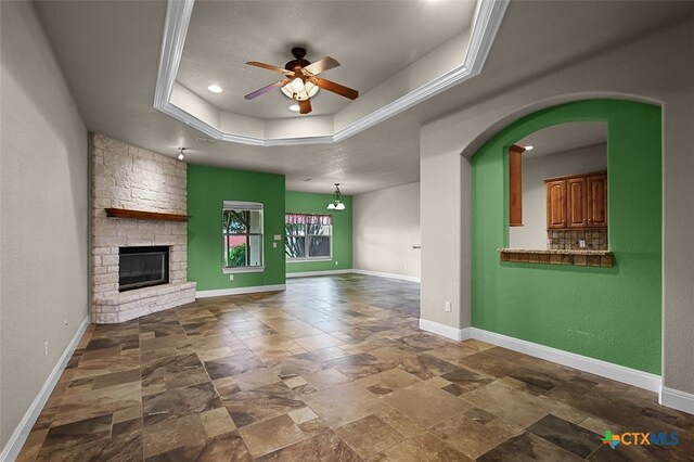 unfurnished living room with a stone fireplace, ornamental molding, ceiling fan, and a tray ceiling