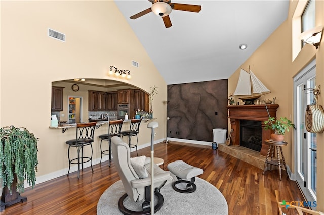 living room featuring high vaulted ceiling, a ceiling fan, dark wood-style flooring, a fireplace with raised hearth, and baseboards