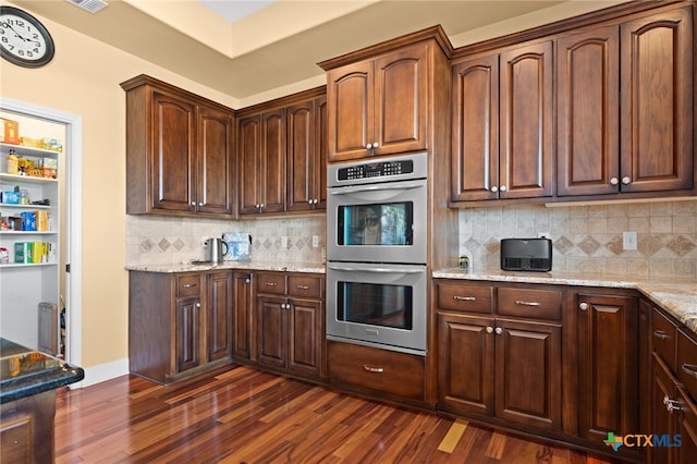 kitchen with double oven, dark wood finished floors, light stone countertops, and decorative backsplash