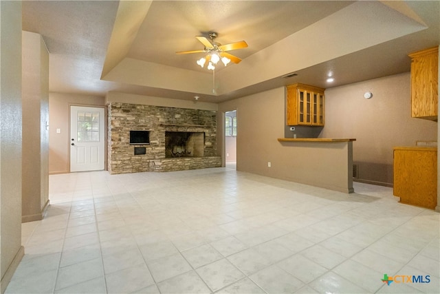 unfurnished living room with light tile patterned floors, ceiling fan, a raised ceiling, and a stone fireplace