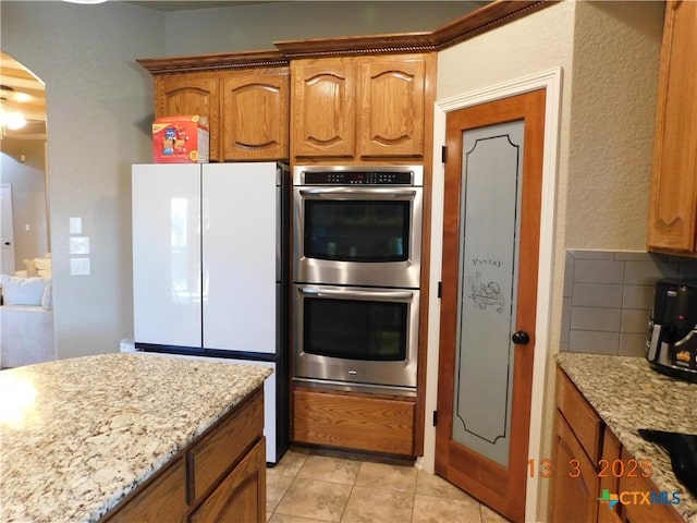 kitchen with light stone counters, tasteful backsplash, freestanding refrigerator, stainless steel double oven, and arched walkways