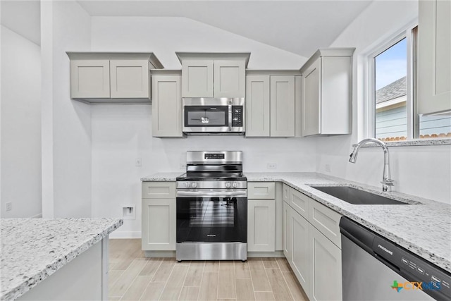 kitchen with gray cabinetry, sink, vaulted ceiling, light stone countertops, and stainless steel appliances