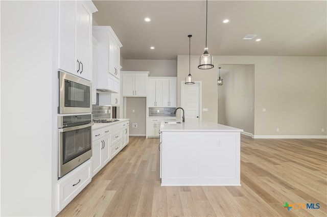 kitchen with white cabinets, a kitchen island with sink, pendant lighting, light wood-type flooring, and appliances with stainless steel finishes