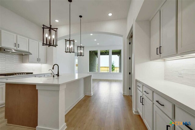 kitchen featuring decorative light fixtures, light hardwood / wood-style floors, white cabinets, stainless steel gas cooktop, and a kitchen island with sink