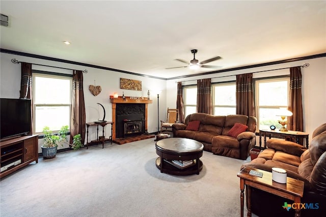living area featuring a fireplace with raised hearth, a healthy amount of sunlight, crown molding, light colored carpet, and ceiling fan