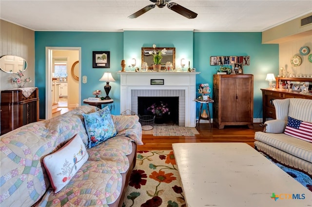 living area with visible vents, a brick fireplace, crown molding, wood finished floors, and a ceiling fan