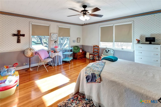 bedroom with a ceiling fan, wood finished floors, a wainscoted wall, wallpapered walls, and ornamental molding