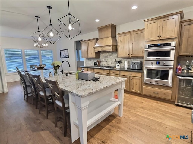 kitchen featuring stainless steel appliances, light stone counters, decorative light fixtures, a center island with sink, and custom exhaust hood