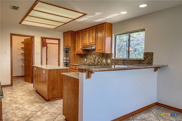 2721 Barton Hill Dr, Bulverde, TX 78163 - kitchen featuring under cabinet range hood, stone countertops, backsplash, brown cabinets, and a center island