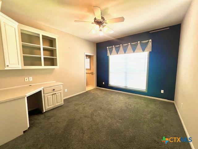 interior space with built in desk, ceiling fan, dark colored carpet, and baseboards