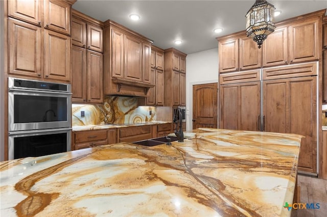 kitchen featuring paneled fridge, hanging light fixtures, light stone countertops, double oven, and sink