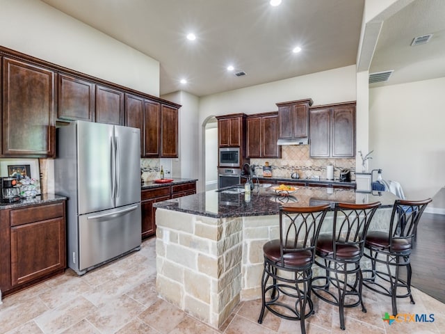 kitchen with dark stone countertops, tasteful backsplash, appliances with stainless steel finishes, and an island with sink