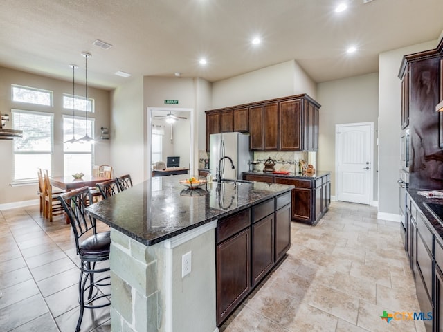 kitchen with dark stone counters, decorative backsplash, an island with sink, ceiling fan, and stainless steel fridge