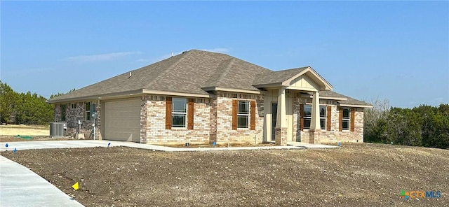 ranch-style home with central air condition unit, an attached garage, driveway, brick siding, and a shingled roof