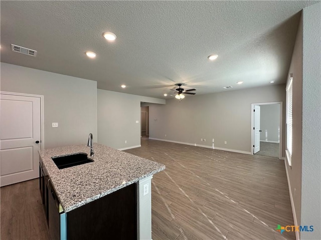 kitchen with a center island with sink, sink, ceiling fan, a textured ceiling, and light stone counters