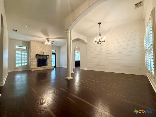 unfurnished living room featuring ceiling fan with notable chandelier, wood walls, a stone fireplace, and dark wood-type flooring